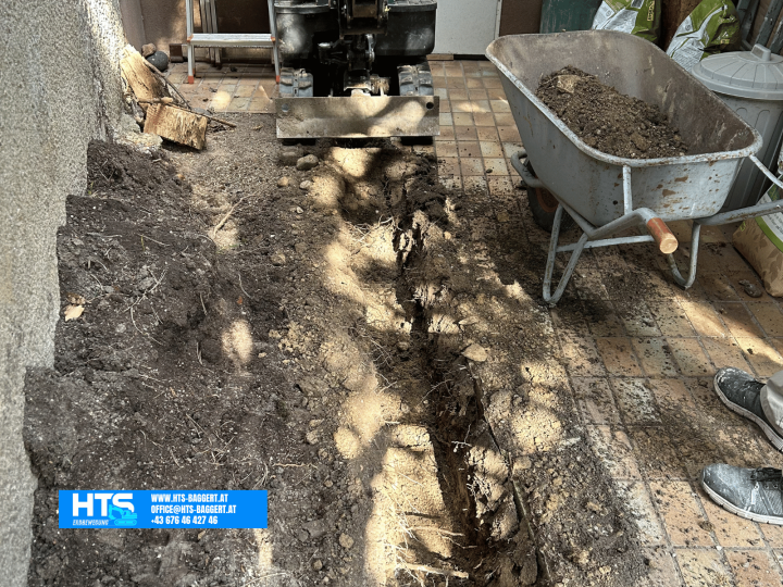 Minibagger von HTS Erdbewegung beim Ausheben einer schmalen Künette in einem engen Gartenbereich zwischen Haus und bestehender Pflasterfläche.