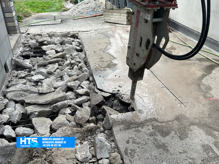 Hydraulikhammer beim Öffnen einer stark bewehrten Betonfläche mit sichtbarem Bewehrungsstahl neben einem bestehenden Gebäude.