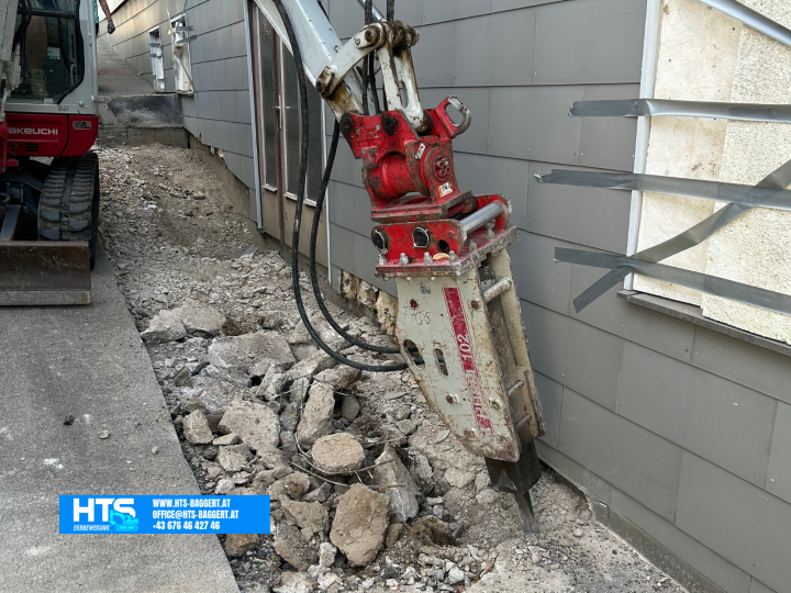 Minibagger von HTS Erdbewegung mit Hydraulikhammer beim Öffnen einer massiven Betonfläche direkt entlang der Hauswand für die spätere Trockenlegung.