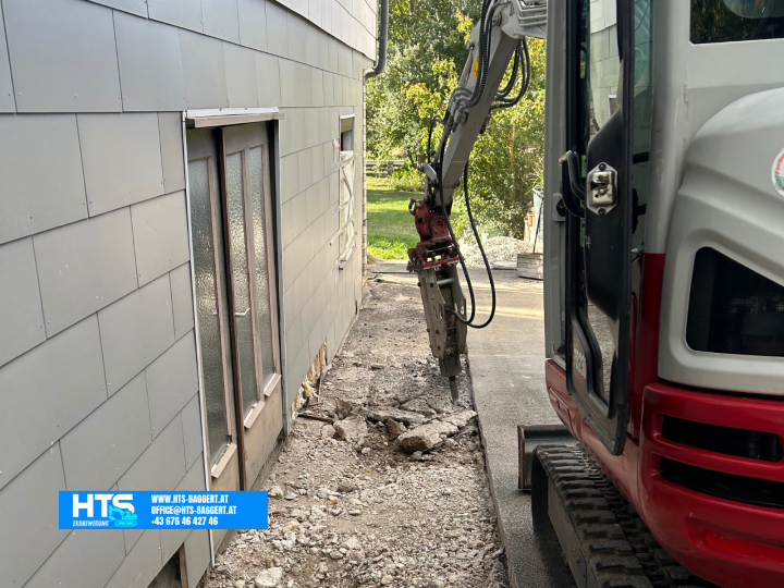 Minibagger mit Hydraulikhammer bei Arbeiten auf engem Raum direkt zwischen Hauswand und bestehender befestigter Fläche.