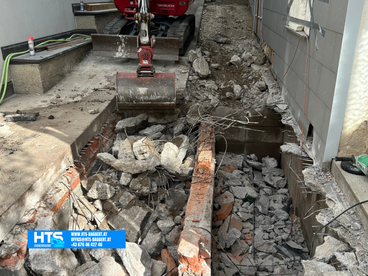 Freigelegter Schachtbereich mit Betonstücken, Mauerwerk und Bewehrungsstahl während der vorbereitenden Arbeiten entlang des Gebäudes.