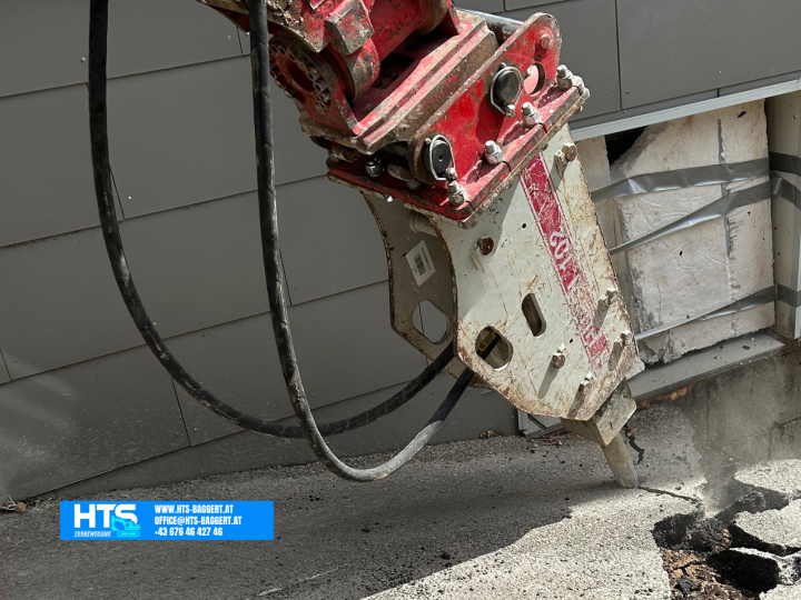 Hydraulikhammer am Minibagger beim Öffnen einer befestigten Betonfläche direkt neben dem Gebäude zur Vorbereitung der weiteren Erdarbeiten für die Trockenlegung.