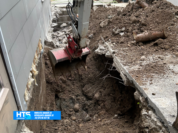 Minibagger beim Aushub entlang einer bestehenden Hauswand auf engem Raum zur Vorbereitung der späteren Trockenlegung und Drainage des Gebäudes.